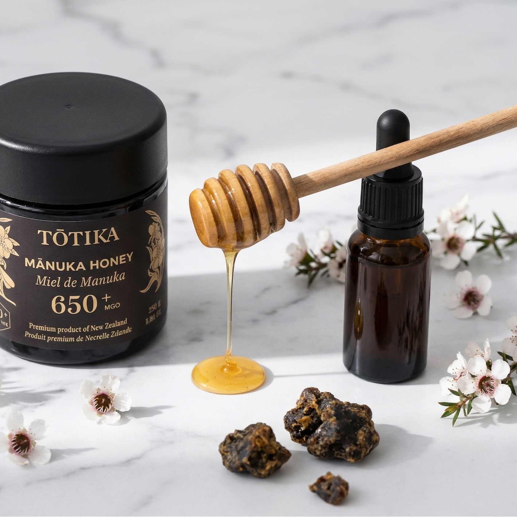 Tōtika mānuka honey jar and propolis dropper bottle side by side on white marble with wooden honey dipper and mānuka blossoms, New Zealand bee products.